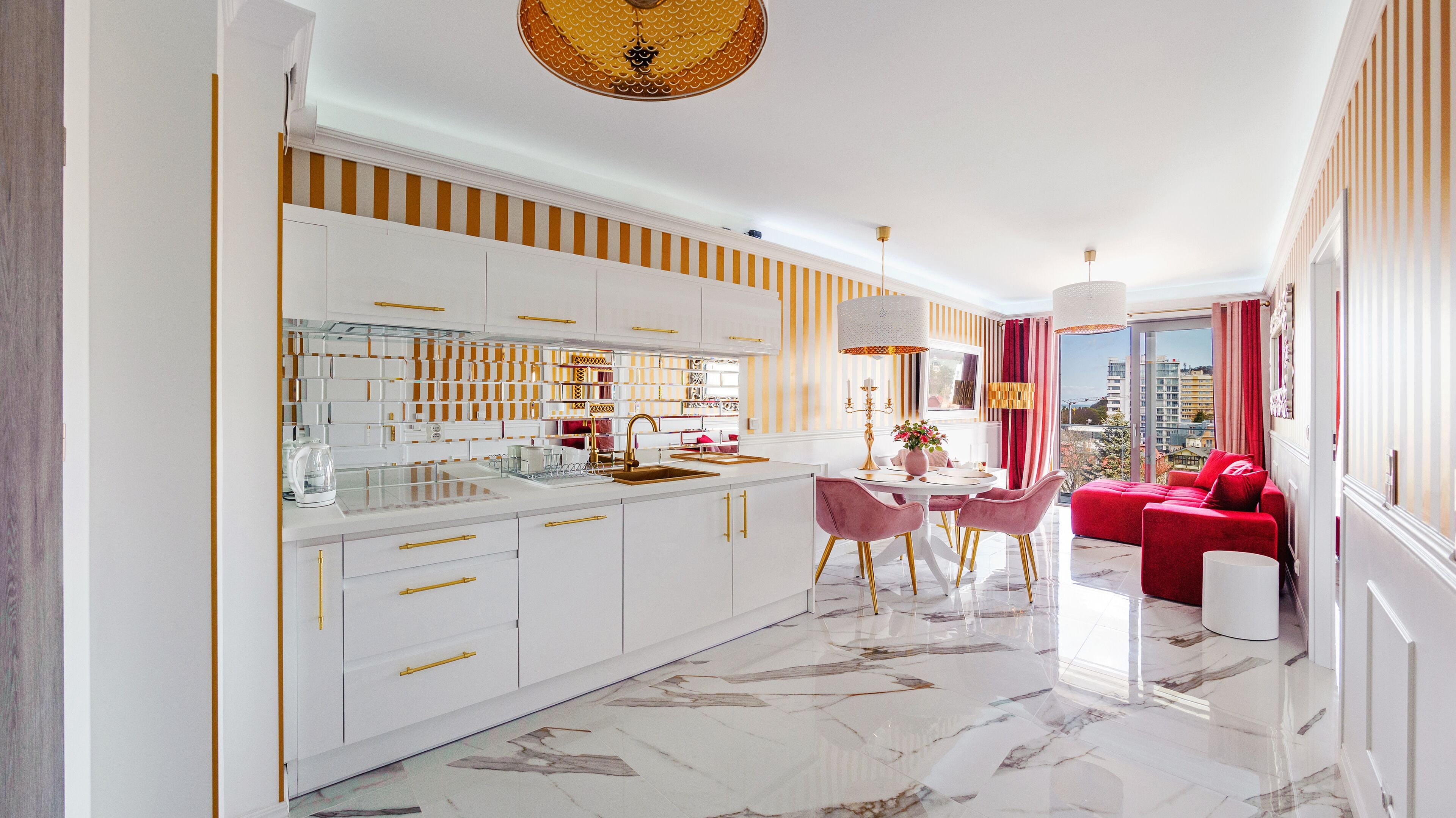 A modern kitchen with white cabinets and gold handles, a dining area with a round table and chairs, and a red sofa in a living room with a view of the city.
