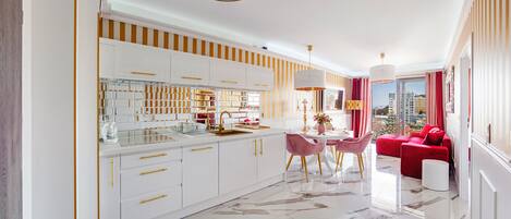 A modern kitchen with white cabinets and gold handles, a dining area with a round table and chairs, and a red sofa in a living room with a view of the city.