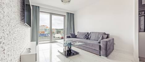 A modern living room with a glass coffee table, grey sofa, and a view of buildings through large windows.
