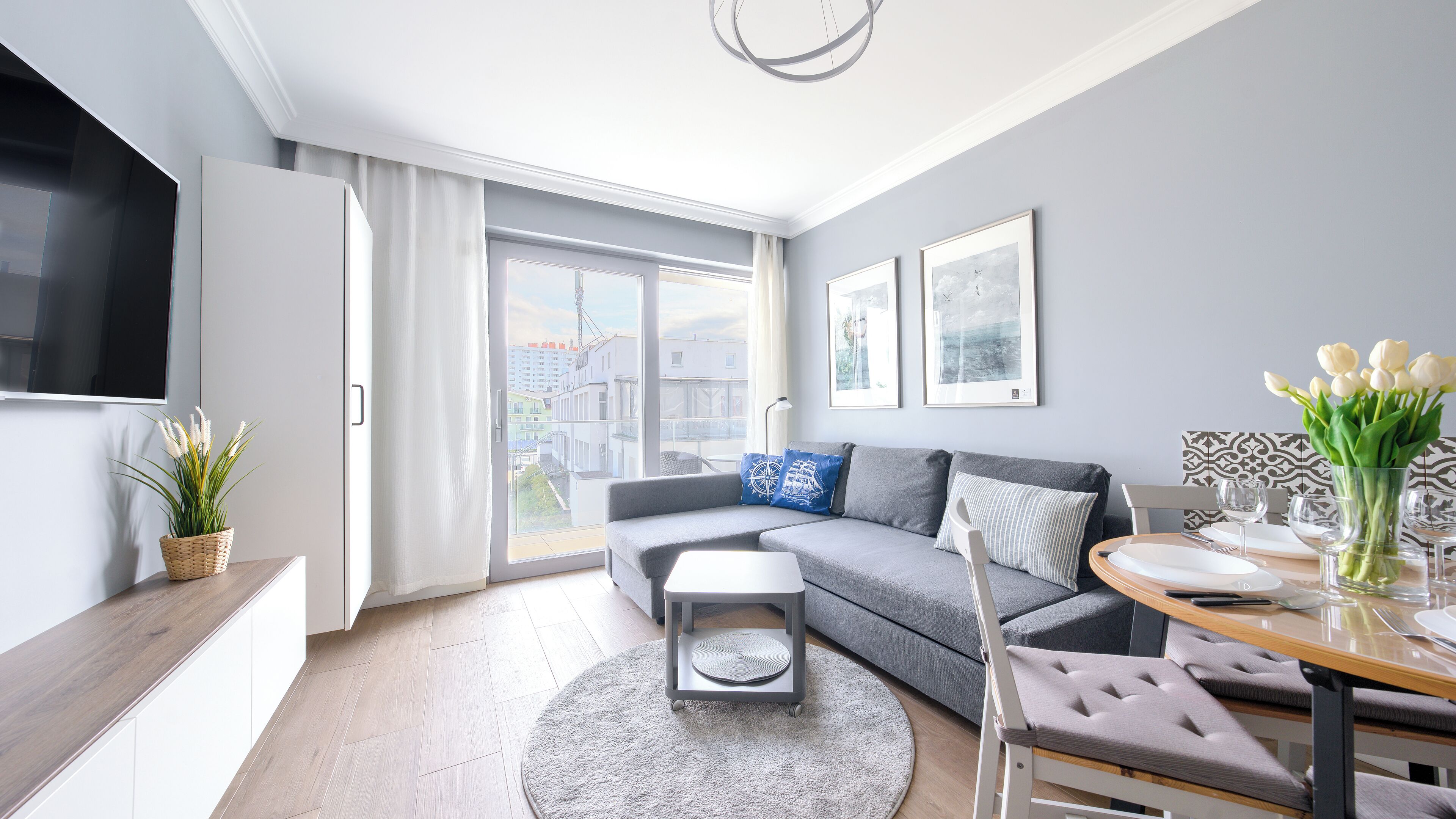 Ein modernes Wohnzimmer mit einem grauen Sofa, einem Esstisch mit Stühlen, einem Flachbildfernseher, einer Topfpflanze und einem großen Fenster mit Blick auf die Stadt.