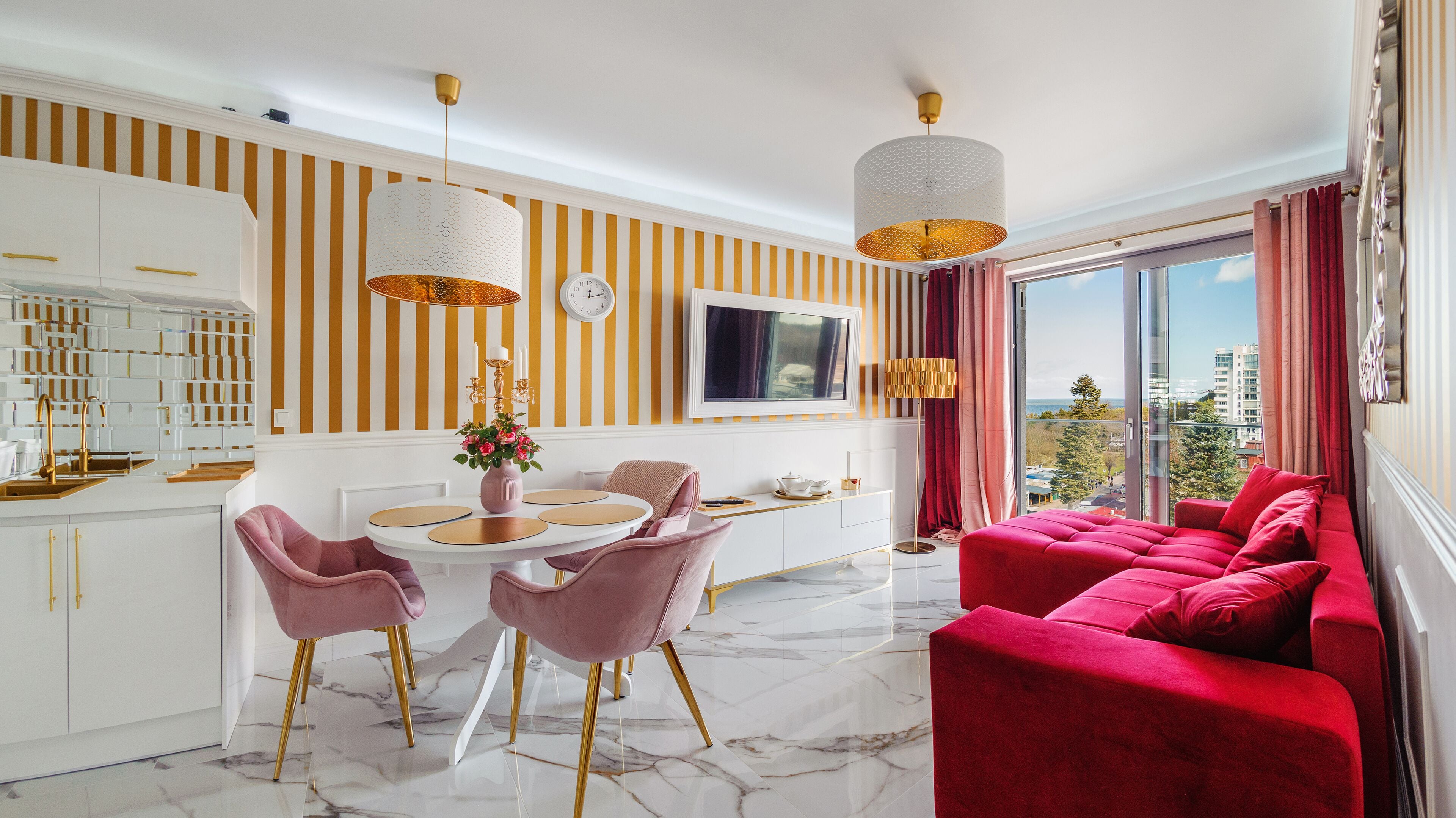 A modern living room with a red sofa, a dining area with pink chairs, a kitchen with white cabinets, and a large window with a city view.