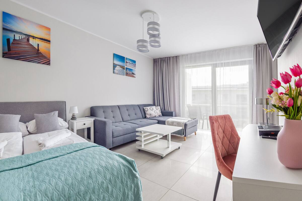 Une chambre d’hôtel moderne avec un lit, un canapé, une table basse, un bureau et une chaise. Il y a un tableau au mur, une fenêtre avec des rideaux et un vase rempli de fleurs sur le bureau.