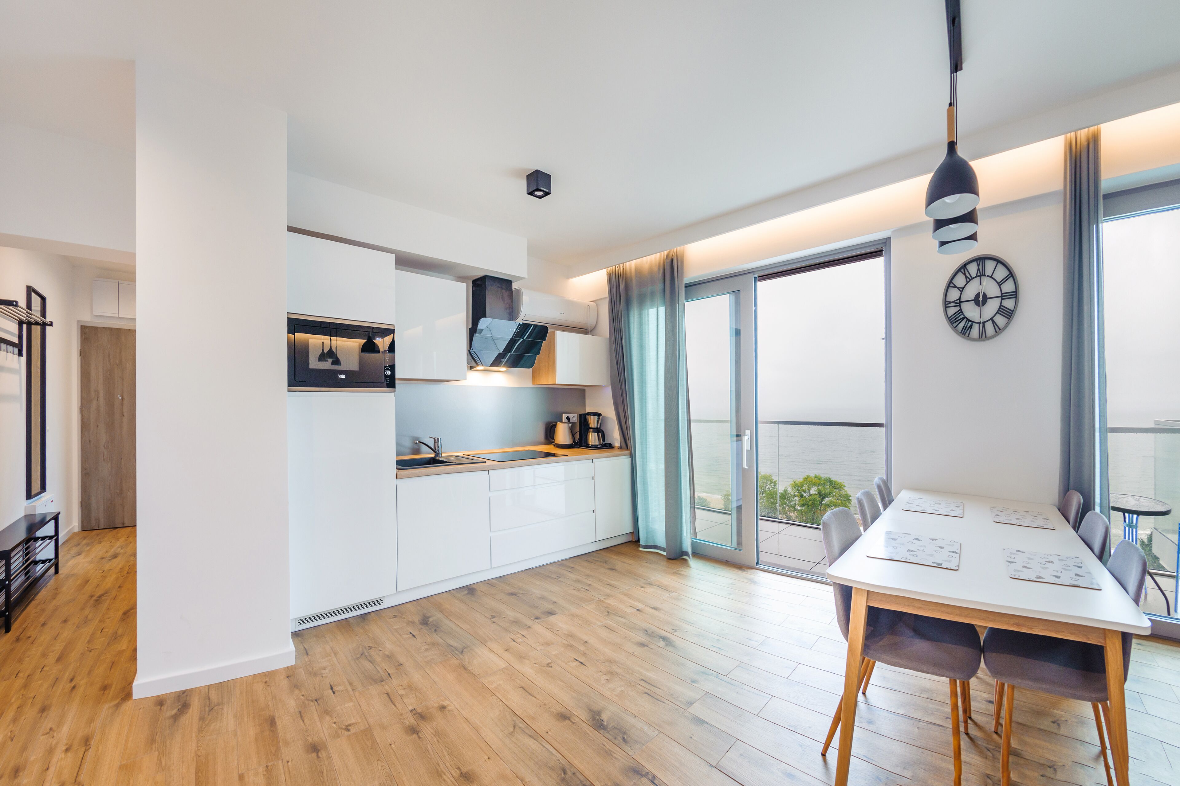 Een moderne keuken met witte kastjes, een houten aanrechtblad en een ingebouwde oven. Er is een eethoek met een tafel en stoelen, en een groot raam met gordijnen.