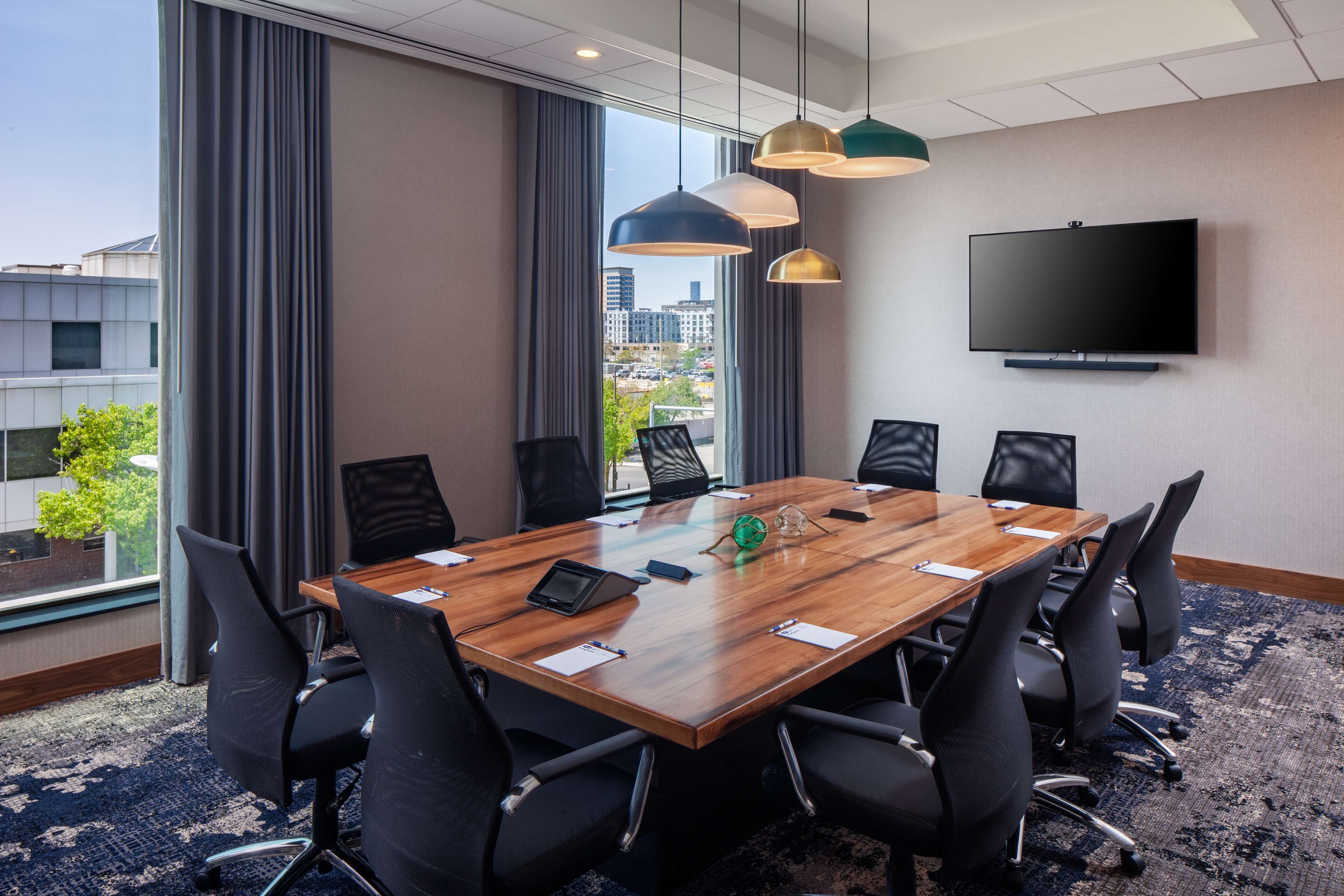 A conference room with a large wooden table, black chairs, a flat-screen TV, and a city view through large windows.