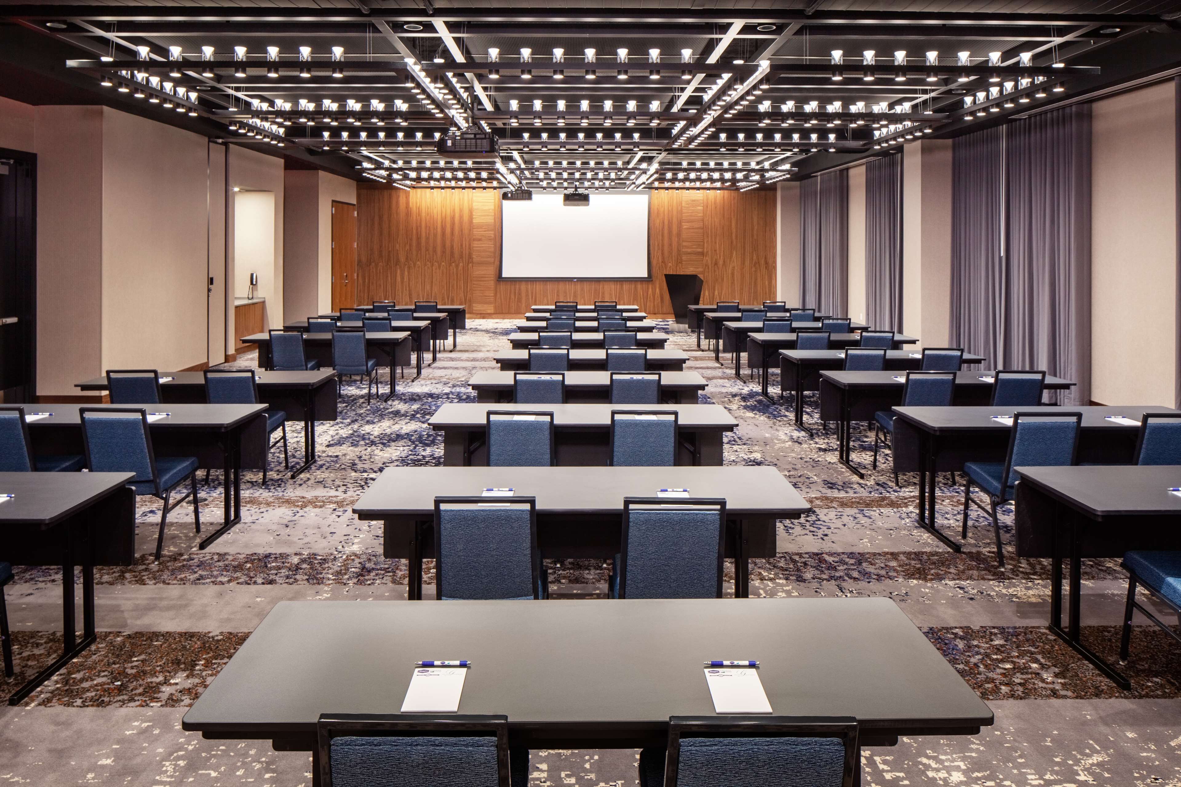 Un salón de conferencias con filas de mesas y sillas, una pantalla grande al frente y suelo alfombrado.