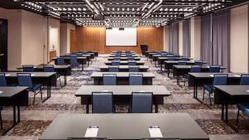 A conference room with rows of tables and chairs, a large screen at the front, and a carpeted floor.