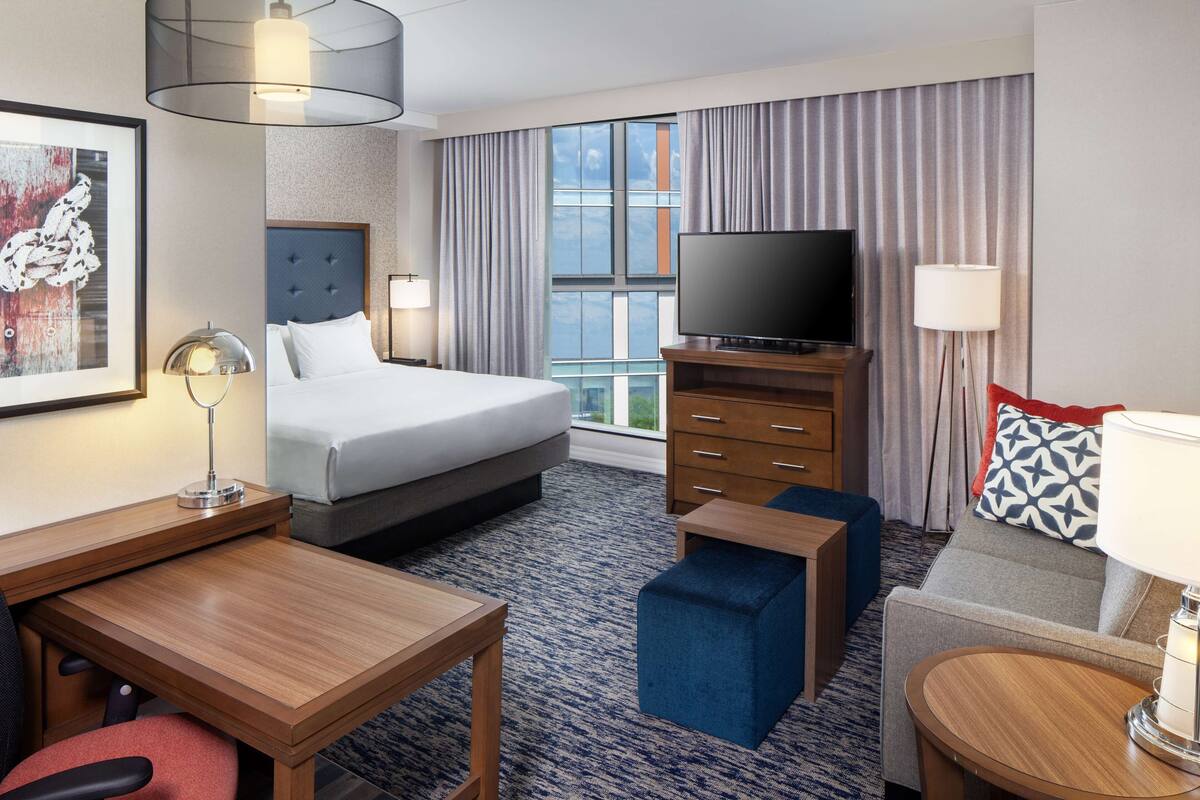 Ein Hotelzimmer mit einem Bett, einem Schreibtisch, einem Fernseher, einer Kommode und einem Fenster mit Vorhängen.