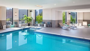 A modern indoor swimming pool with clear blue water, sun loungers, and large windows offering views of the city. - Homewood Suites by Hilton Boston Seaport District (Boston)