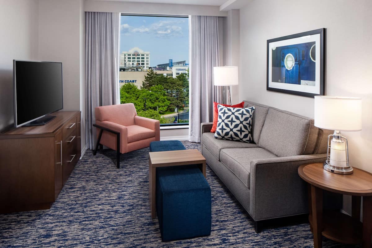 Ein Hotelzimmer mit Fernseher, einer Couch, einem Sessel, einem kleinen Tisch und einer Lampe. Ein Fenster bietet Blick auf Gebäude und Bäume.