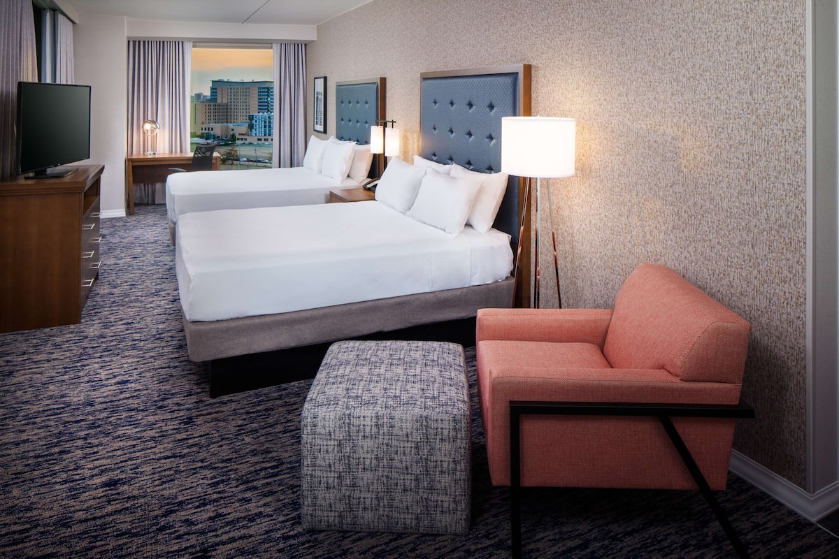 Ein Hotelzimmer mit zwei Betten, einem Fernseher, einem Schreibtisch und Blick auf die Stadt.