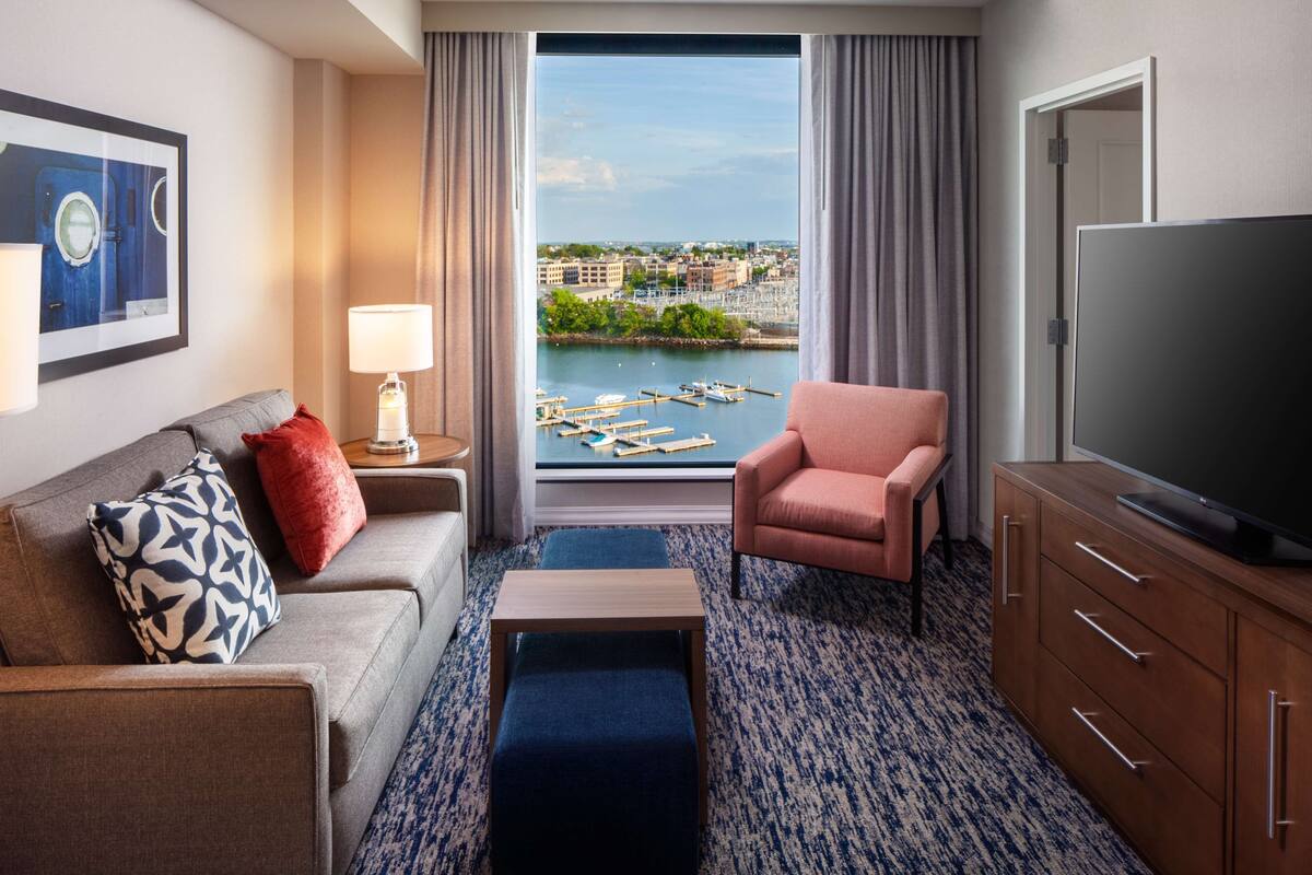 Ein Hotelzimmer mit Sofa, Sessel, Fernseher und Blick auf den Yachthafen.