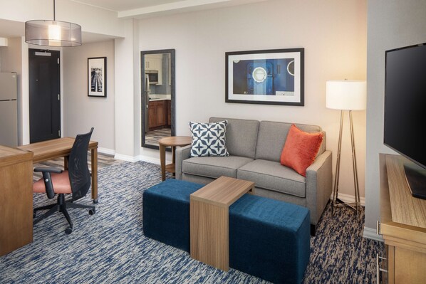 A hotel room with a desk, chair, sofa, ottoman, TV, and a lamp.