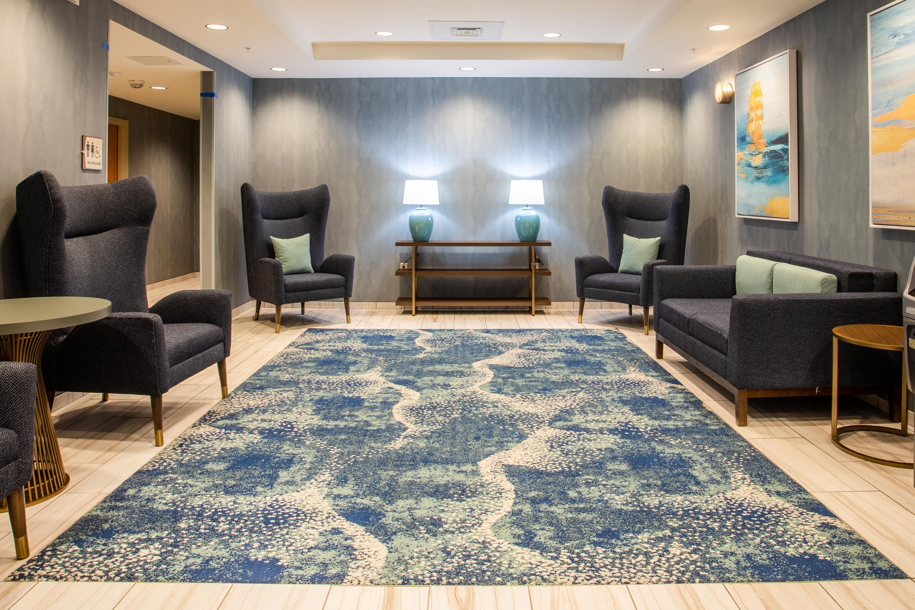 A hotel lobby with a large blue patterned rug, two armchairs, a sofa, a small round table, and a bench.