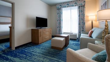 Habitación de hotel con cama, televisión, armario de madera, sofá, sillón y ventana con cortinas.