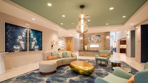 Eine großzügige Lobby mit einer gebogenen Couch, einem runden goldenen Hocker und einem blau gemusterten Teppich. An den Wänden hängen zwei Gemälde, und eine große Chandelier erleuchtet den Raum.