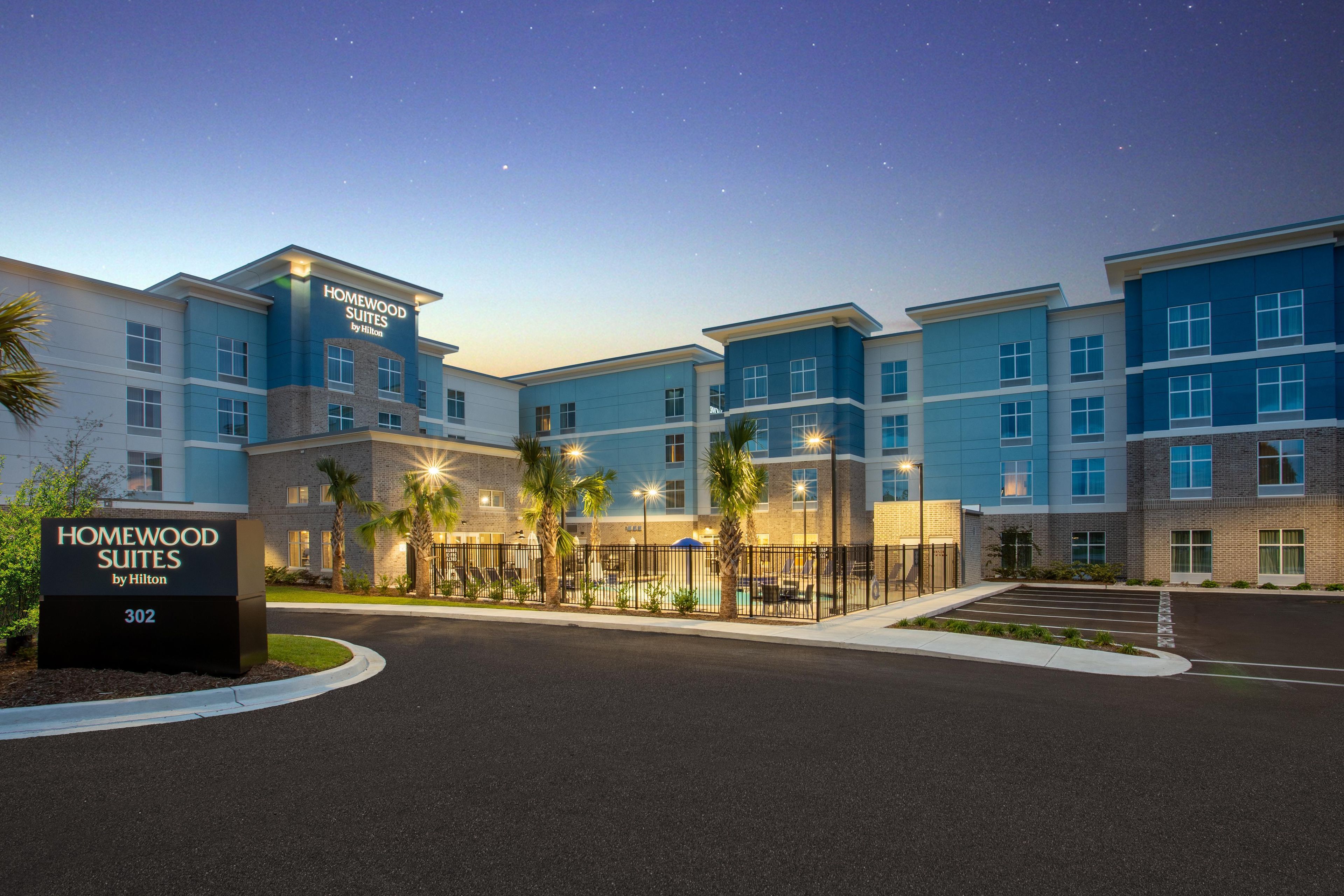 A hotel with multiple-story buildings, a sign indicating 'Homewood Suites by Hilton,' and a parking area with a fence and palm trees.