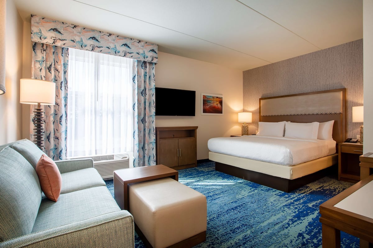 A hotel room with a bed, a sofa, a small table, a nightstand, a TV, and a window with curtains.