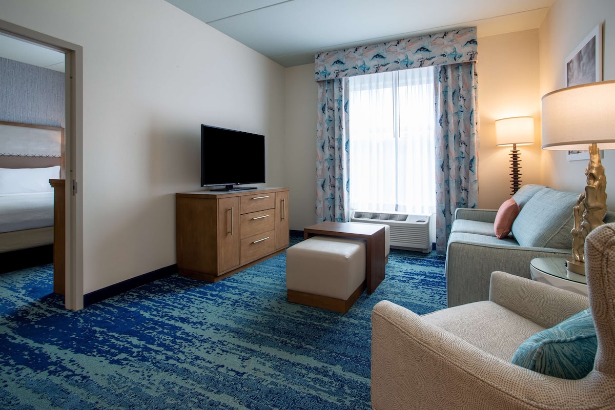 A hotel room with a bed, a TV on a wooden cabinet, a sofa, a chair, a lamp, and a window with curtains.