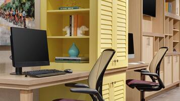 Un ufficio moderno con scrivania, due sedie ergonomiche, un monitor per computer, tastiera e mouse. Si vedono libri e oggetti decorativi sugli scaffali e un quadro appeso al muro.