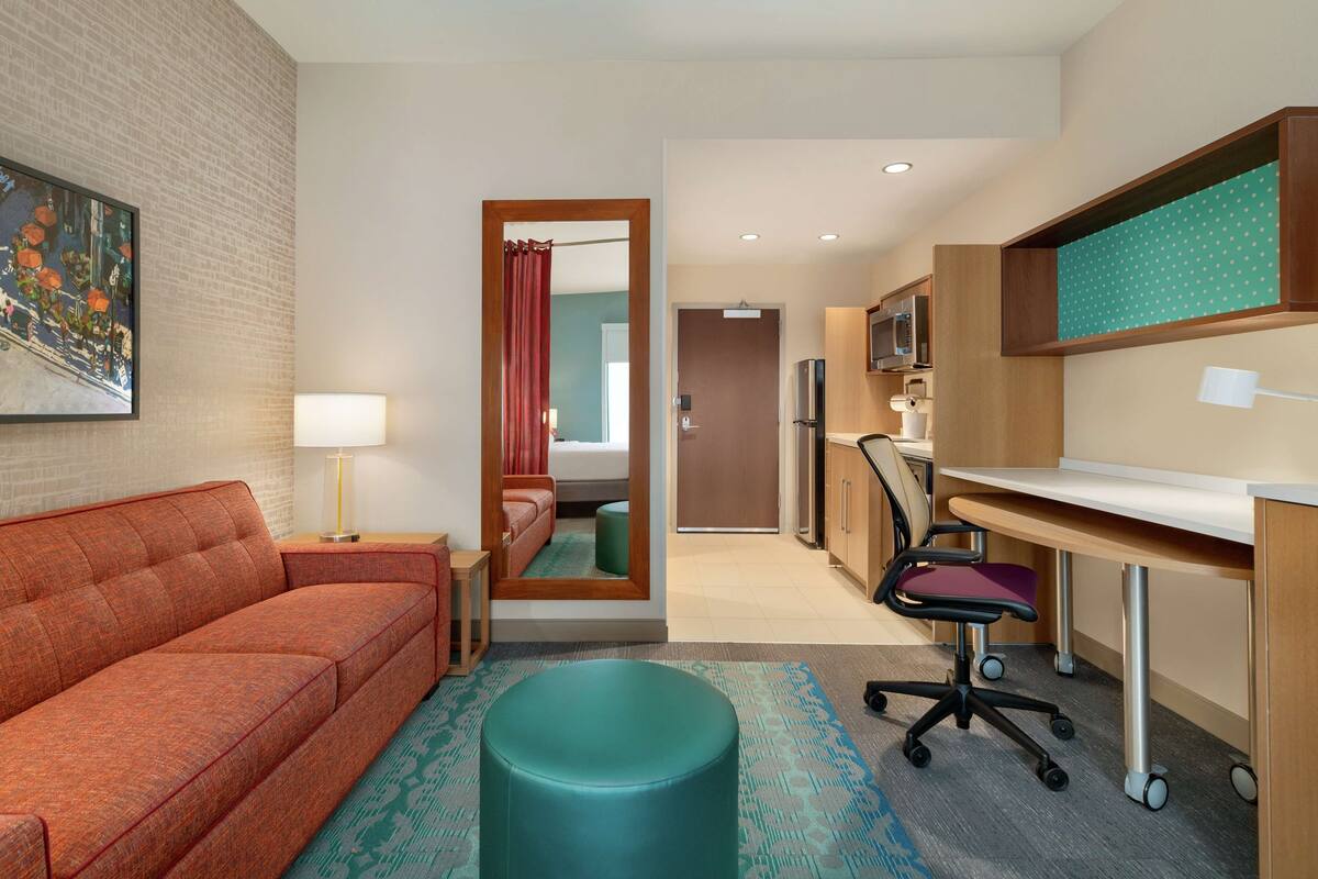 A hotel room with a sofa, a teal ottoman, a desk with a chair, a kitchenette, and a bathroom visible through an open door.