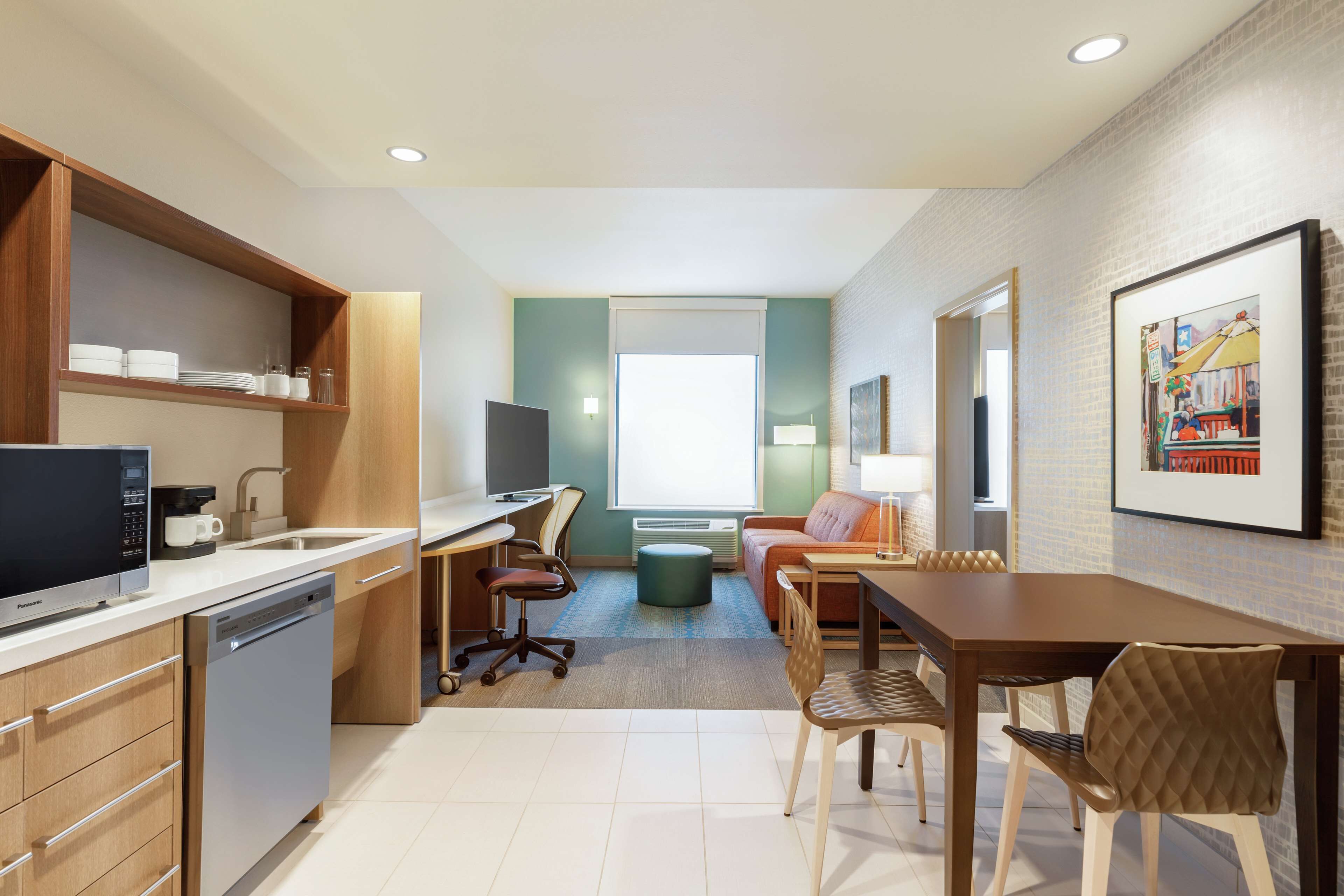 A modern hotel room with a kitchenette, dining area, and a large window providing natural light.