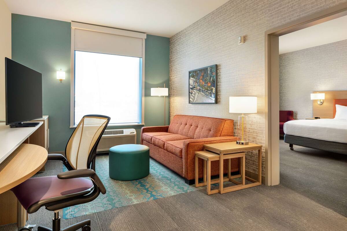 A hotel room with a desk, chair, sofa, bedside table, lamp, bed, and a painting on the wall.
