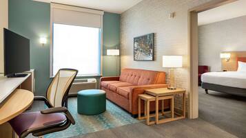 Habitación de hotel con escritorio, silla, sofá, mesita de noche, lámpara, cama y un cuadro en la pared.