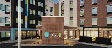 A modern hotel with a prominent "tru HOME 2 by Hilton" sign, featuring a mix of brick and contemporary architecture, and a well-lit entrance area.