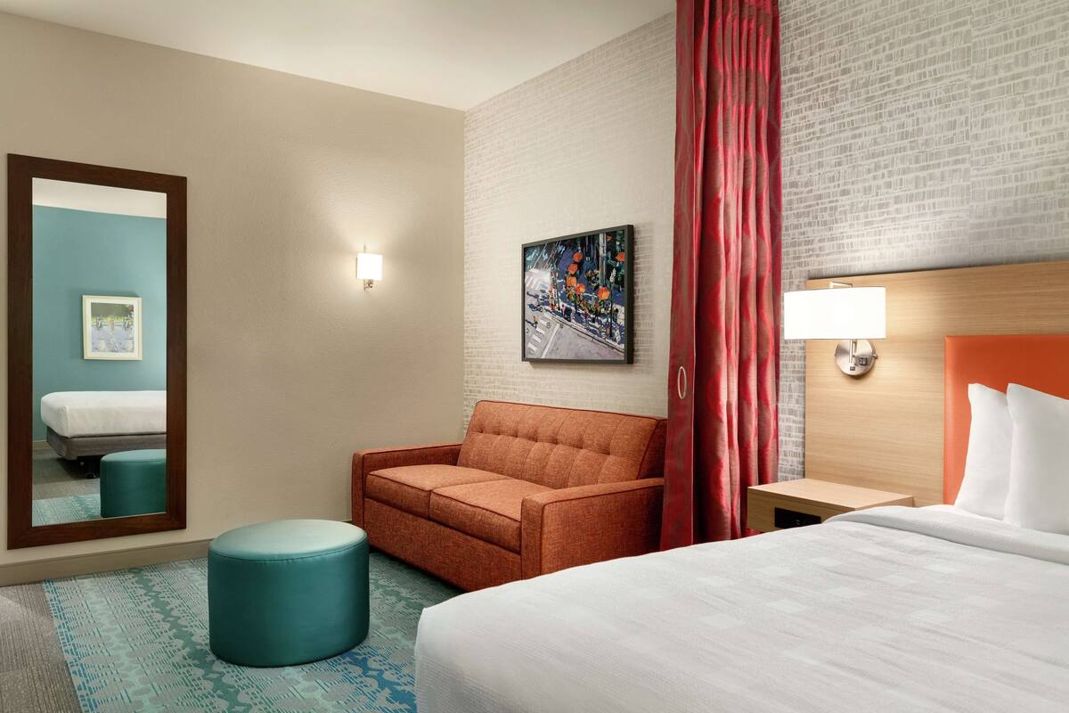A hotel room with a bed, a sofa, a small ottoman, a nightstand, a wall-mounted lamp, a painting, and a mirror.