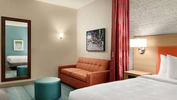 Habitación de hotel con cama, sofá, pequeño reposapiés, mesita de noche, lámpara de pared, un cuadro y un espejo.