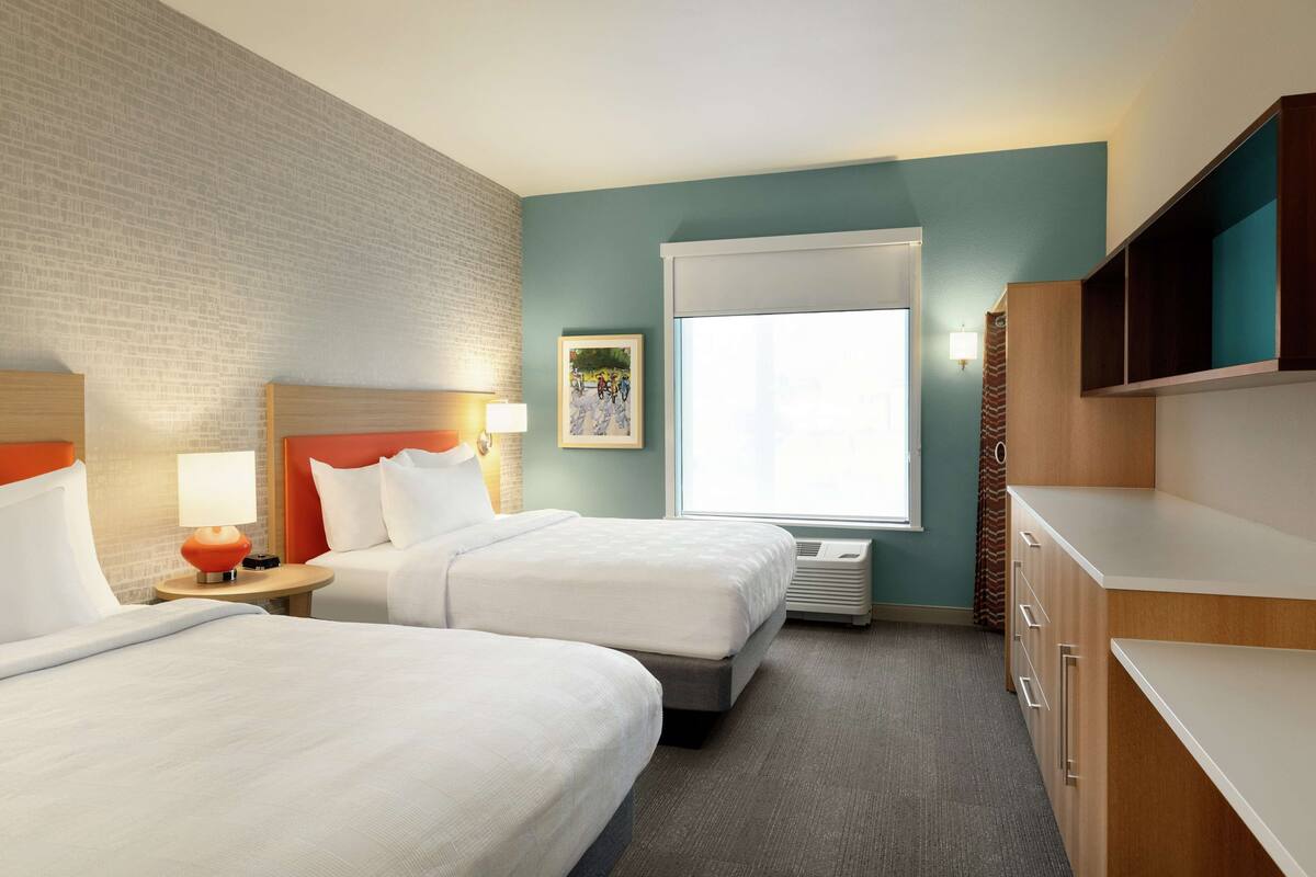 A hotel room with two beds, a large window, a brick accent wall, and a wooden cabinet.