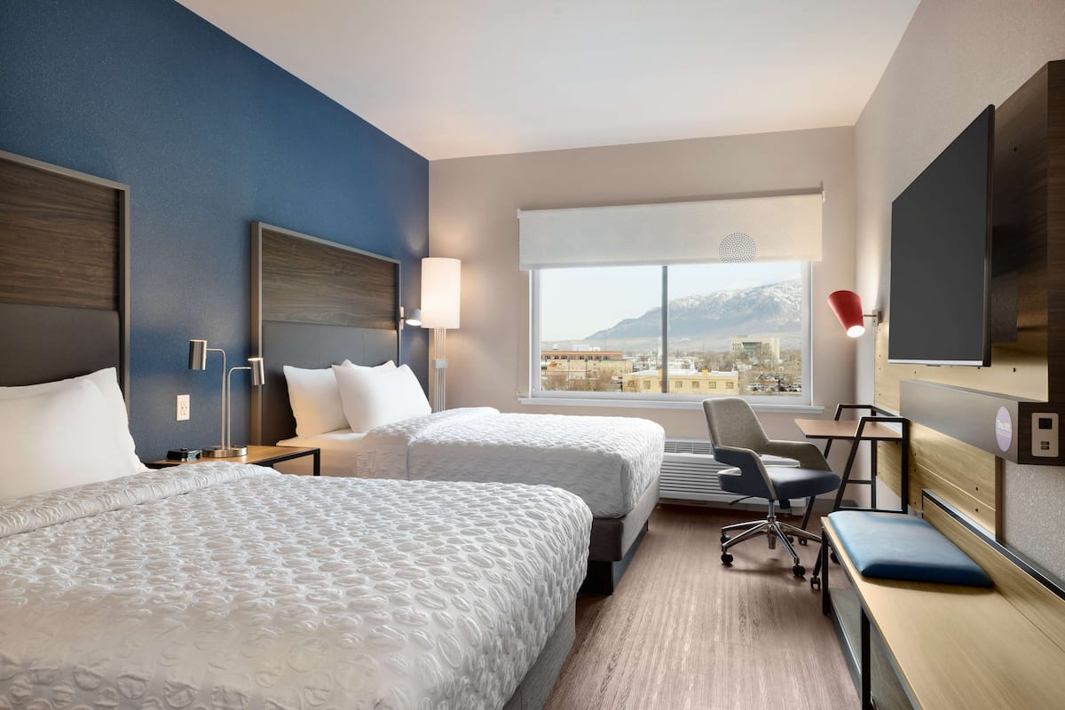 A hotel room with two beds, a desk, a chair, a TV, and a large window with a mountain view.