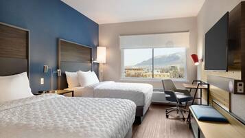 A hotel room with two beds, a desk, a chair, a TV, and a large window with a mountain view.