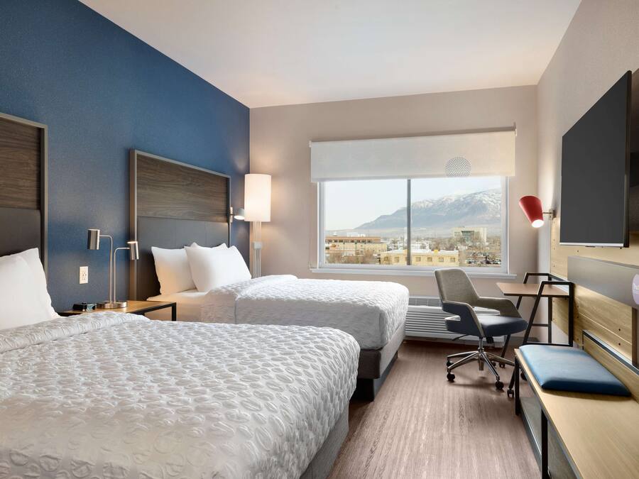 A hotel room with two beds, a desk, a chair, a TV, and a large window with a mountain view.