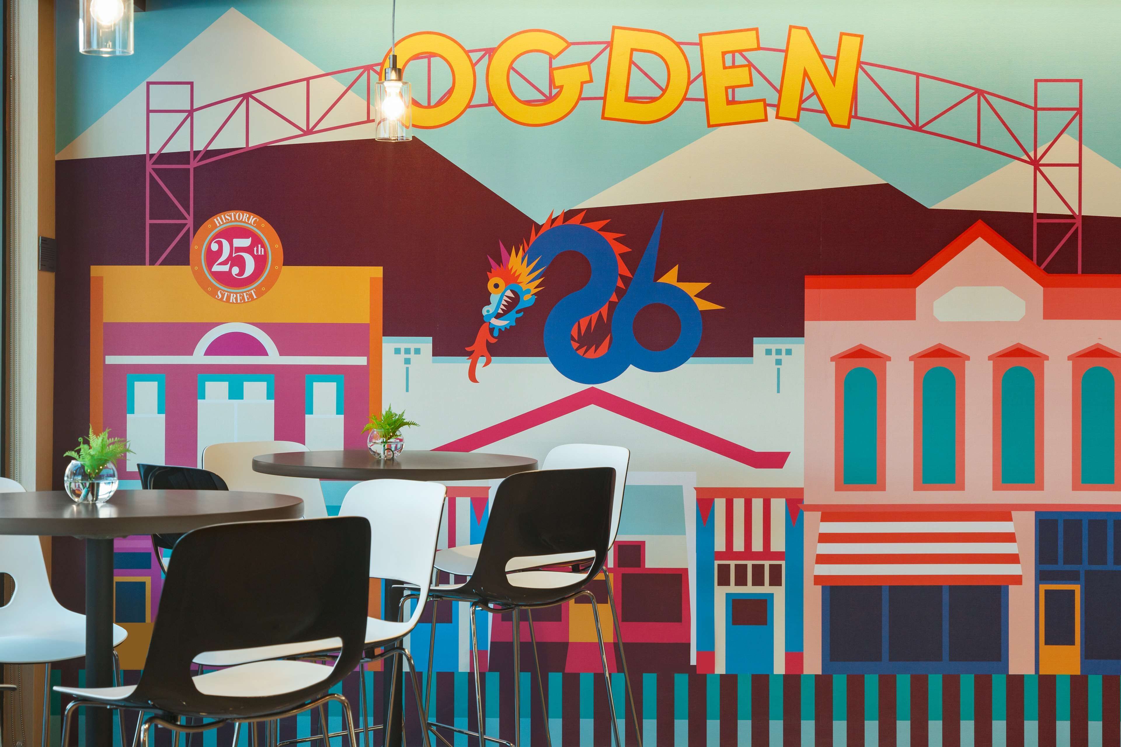 Ein farbenfrohes Wandgemälde mit dem Schriftzug 'OGDEN' und einem Drachen, Tischen, Stühlen und einer Pflanze auf einem Tisch.