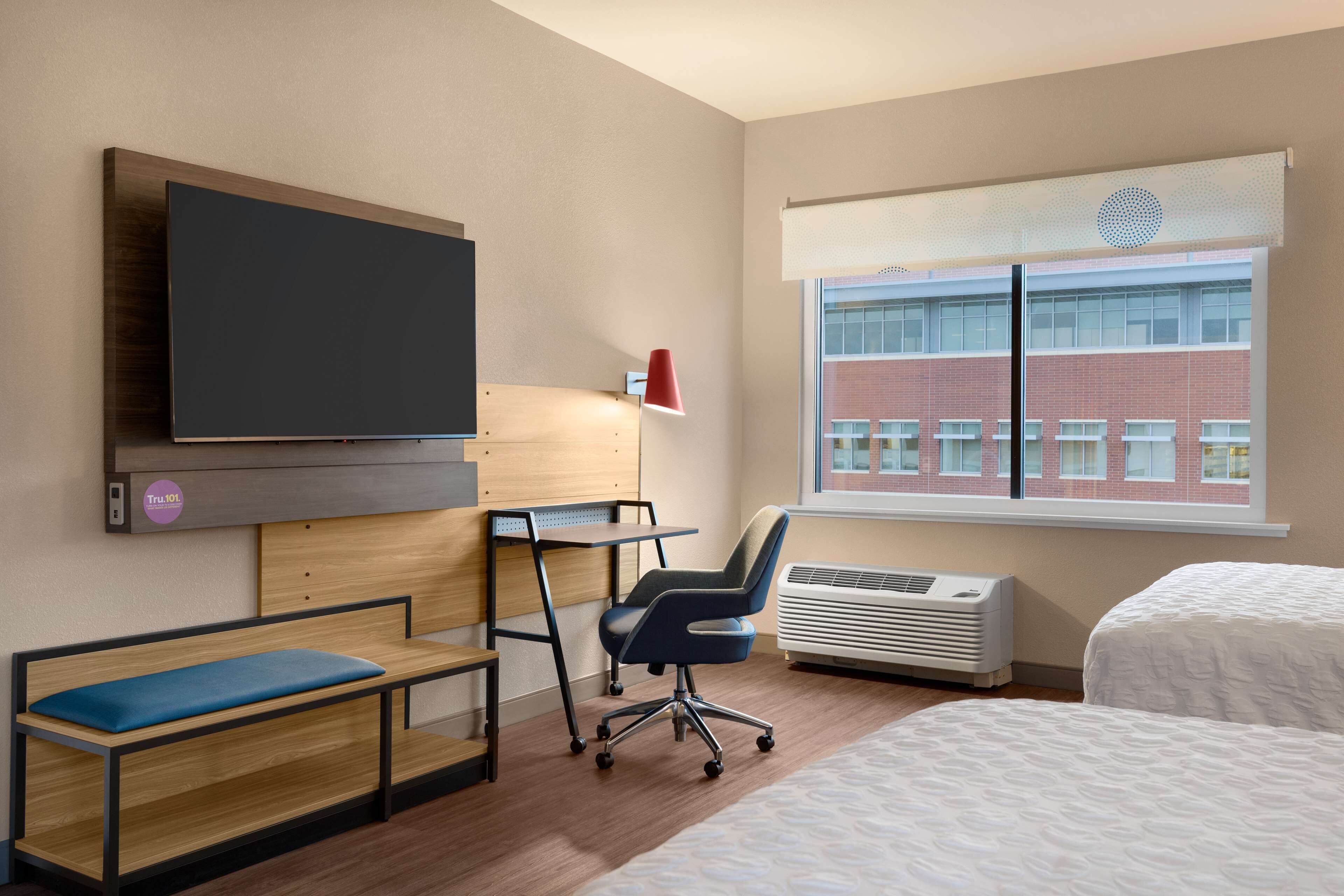 A hotel room with a bed, a desk with a chair, a television mounted on the wall, and a window with a view of a building.