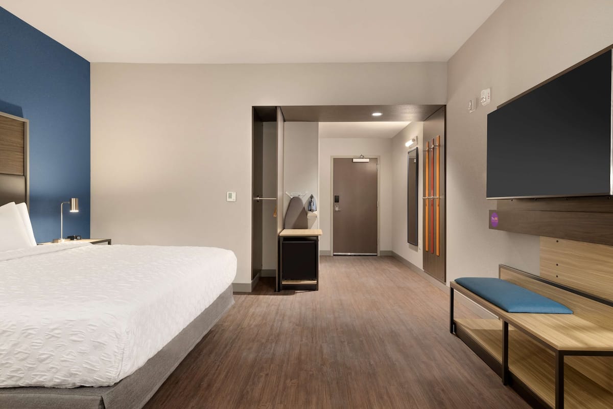 A modern hotel room with a large bed, a desk, a bench, and a TV.