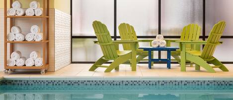A poolside area with a wooden shelf holding rolled towels, a table with chairs, and a view of a building through large windows.