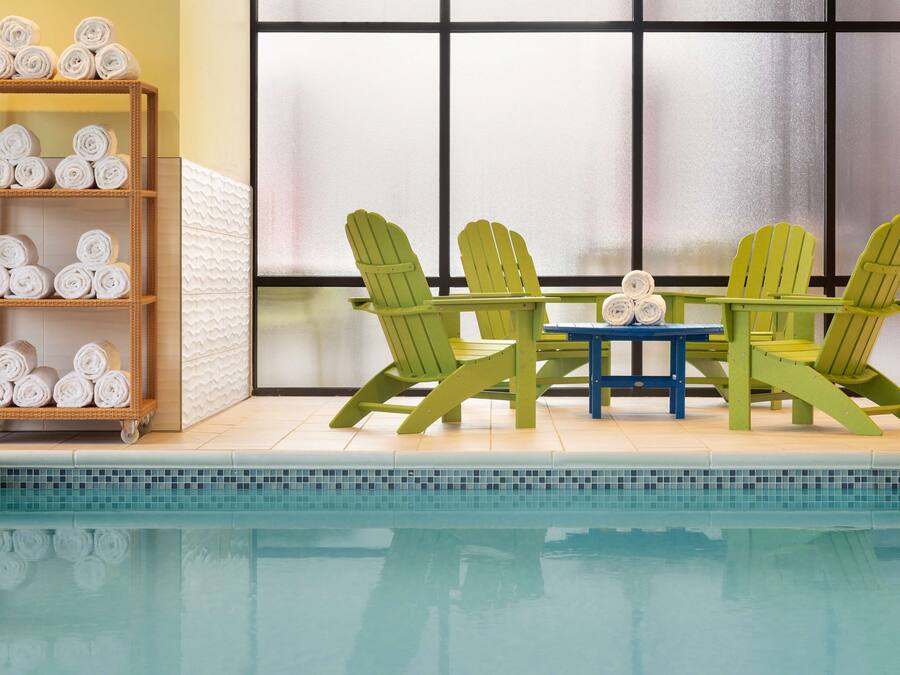 Een zwembadgedeelte met een houten plank met opgerolde handdoeken, een tafel met stoelen en uitzicht op een gebouw door grote ramen.