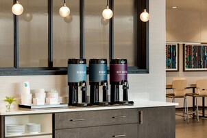 A modern kitchen area with a coffee station featuring three dispensers labeled "Regular," "Decaf," and "Hot Water." There are stacked white cups and a small plant on the counter.