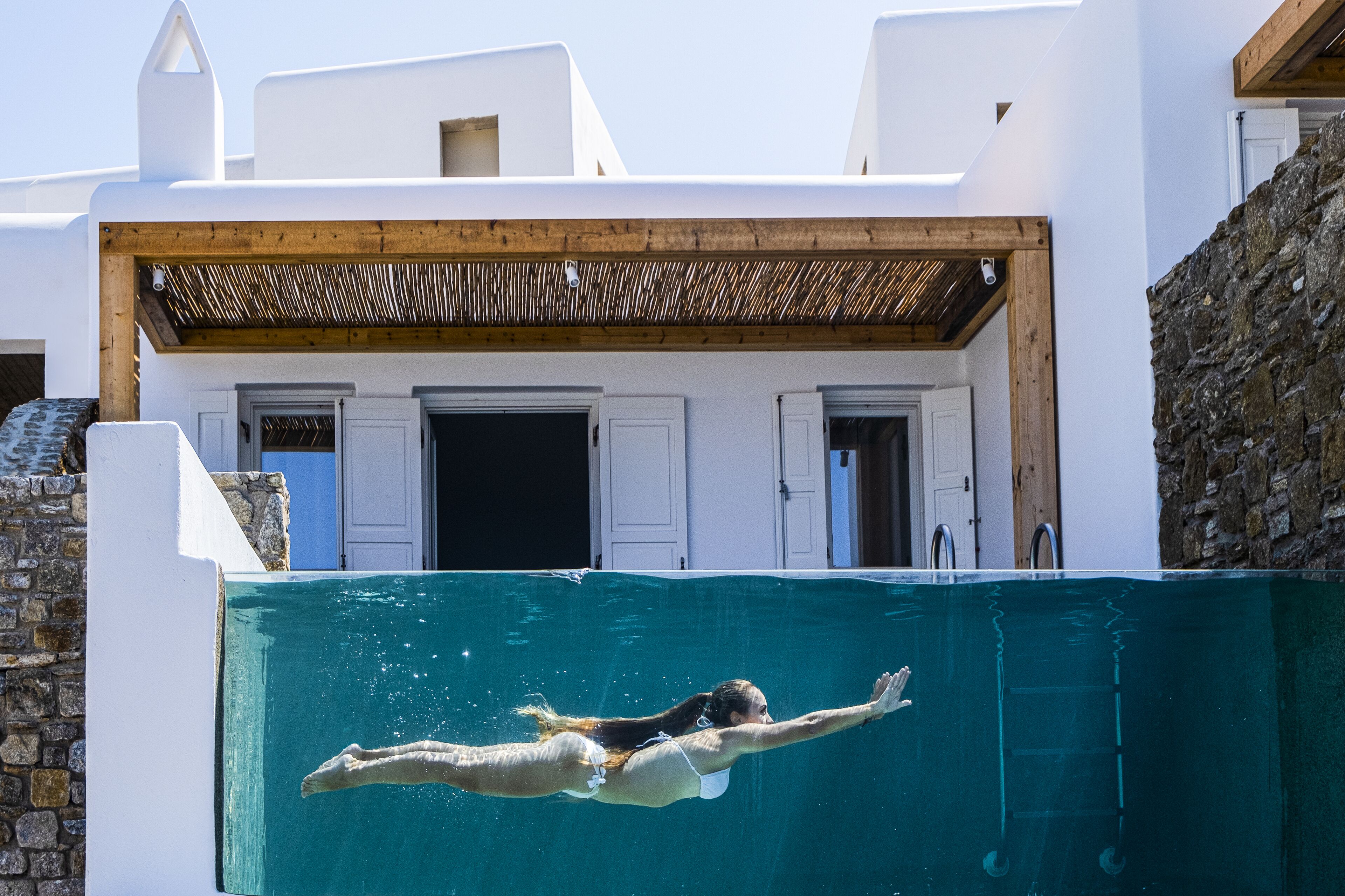 A person swimming in a pool in front of a white building with a wooden ceiling.