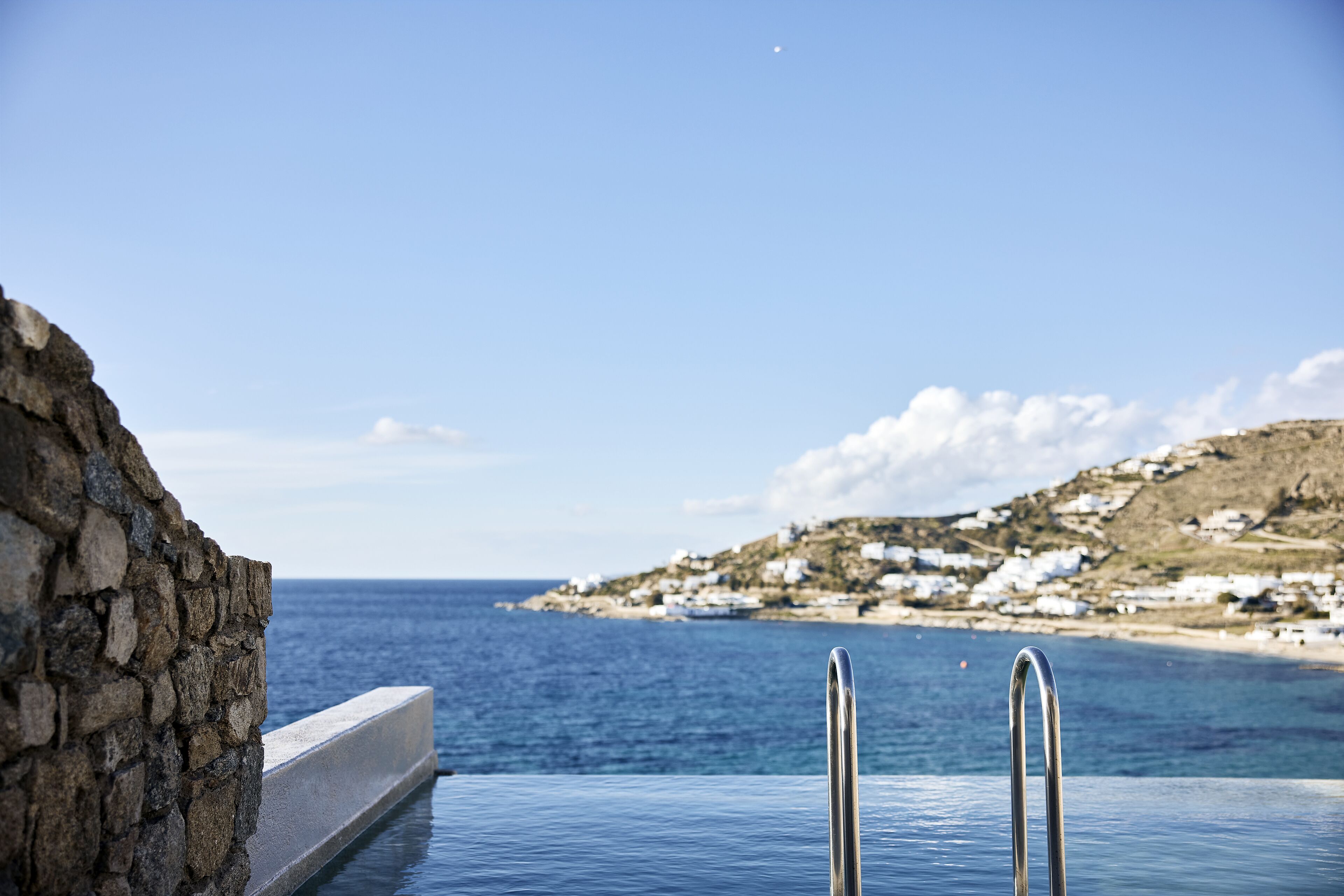 A stone wall and a pool with a metal ladder, overlooking a coastal area with white buildings.