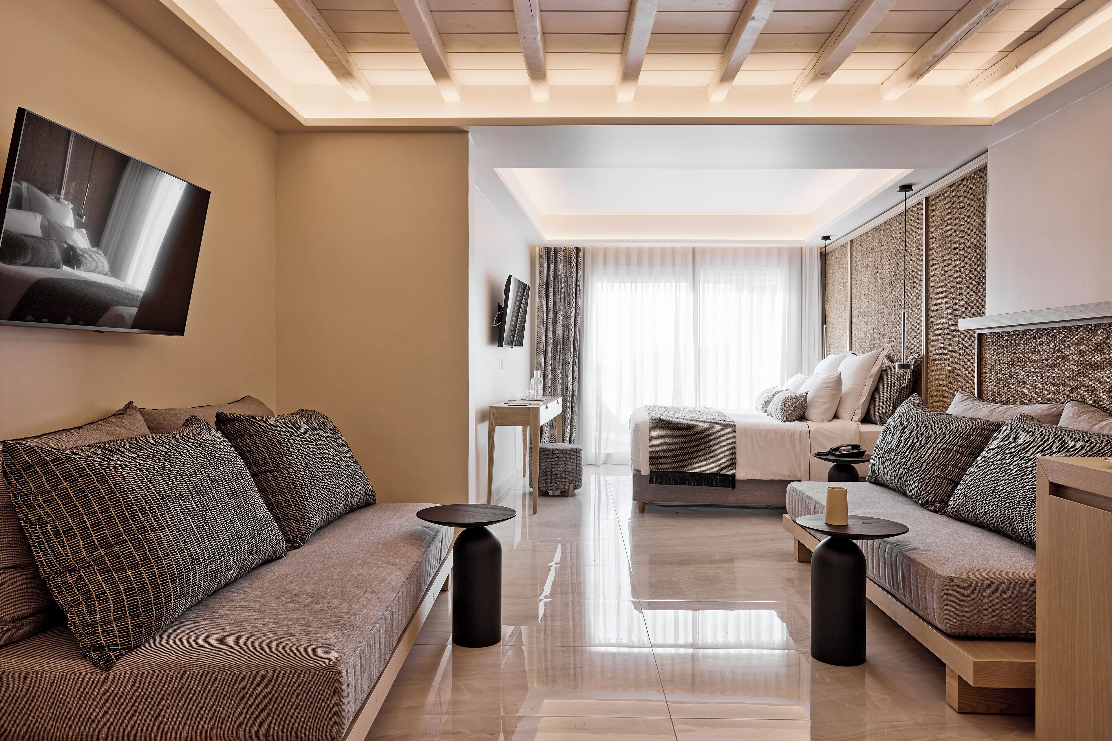 A modern hotel room with a sofa, a bed, a TV mounted on the wall, and a large window with curtains.