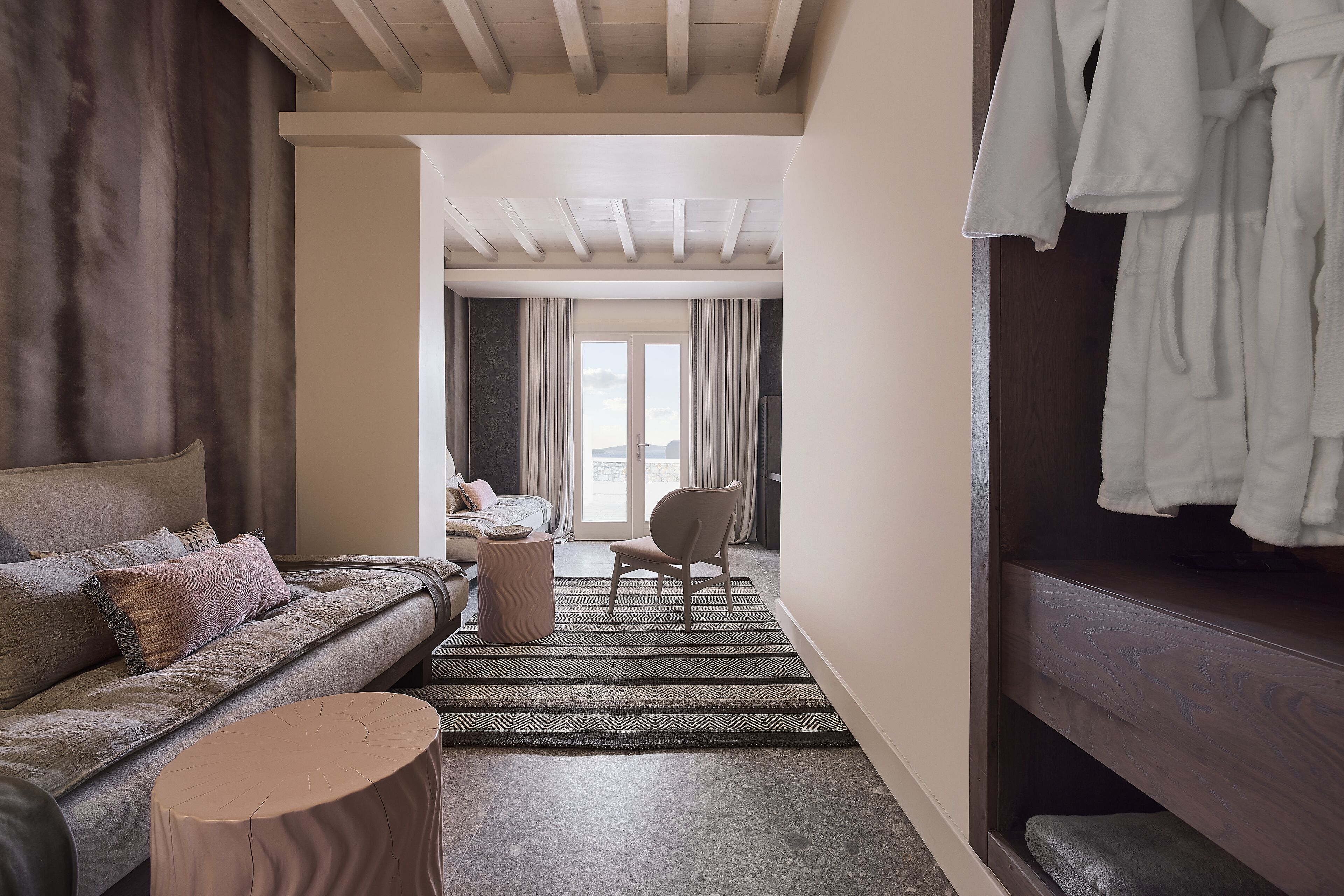 A modern hotel room with a sofa, a chair, a round wooden stool, and a wooden wardrobe with robes.