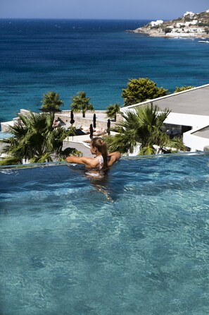 A pool with clear blue water, surrounded by palm trees and a view of the ocean. - Amazon Mykonos Resort & Spa (Mykonos)