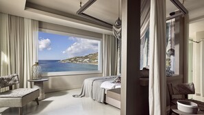 Une chambre avec une grande fenêtre donnant sur un paysage côtier, un lit, une chaise et une petite table sur laquelle est posé un vase.