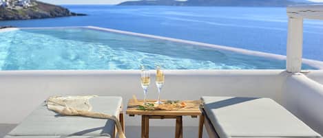 Mesa junto a la piscina con dos copas de vino blanco, un plato de comida y vistas al mar y colinas lejanas.