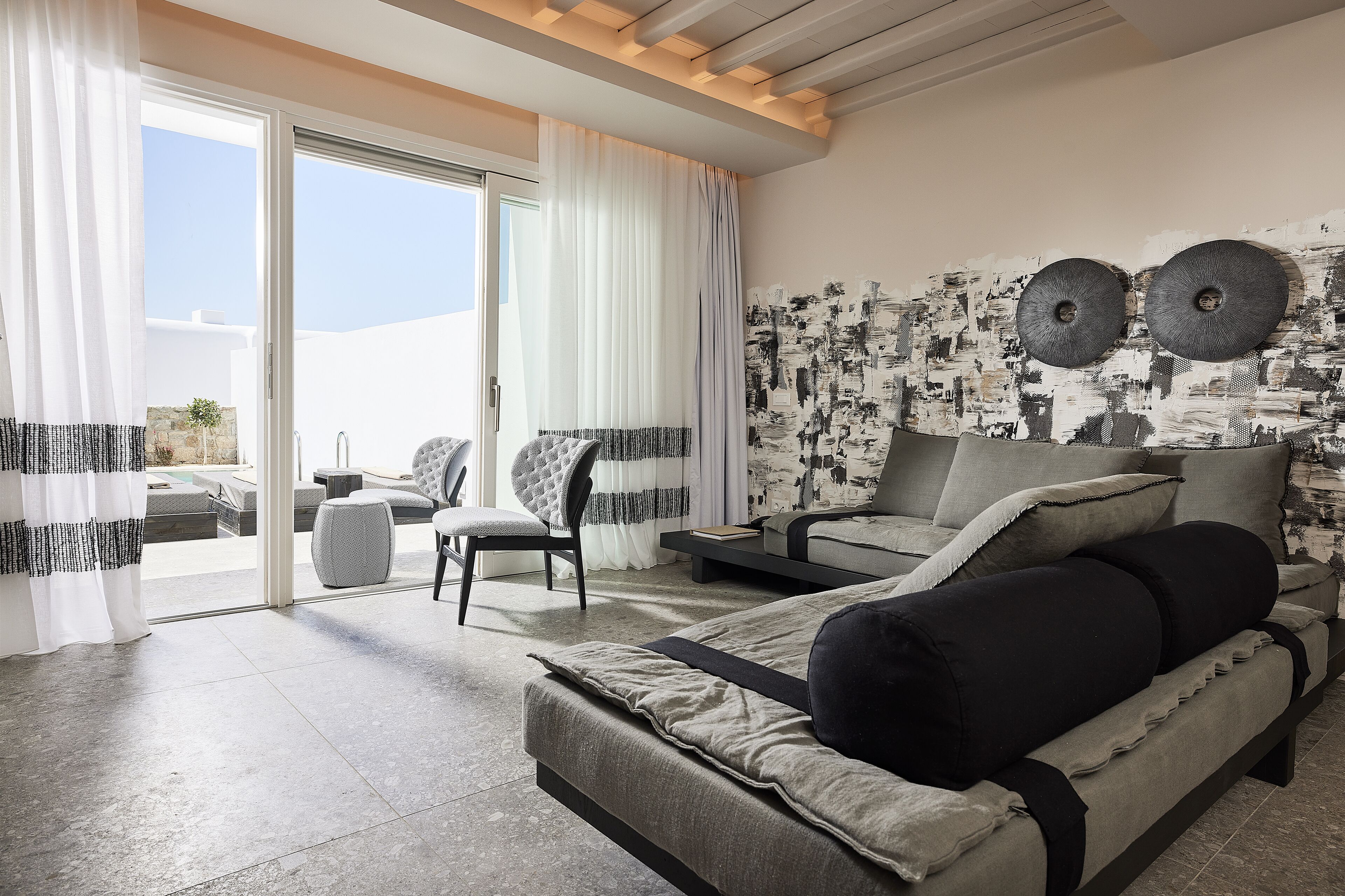 A modern hotel room with a large sliding glass door leading to a balcony, a sofa, a chair, and a bed with a view of the sky.