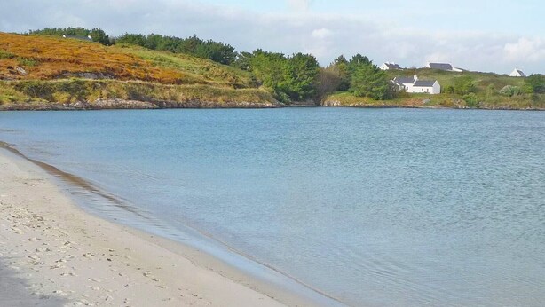 Ein Strand mit sandigem Ufer und ruhigem Wasser. Im Hintergrund befinden sich Häuser auf einem grasbewachsenen Hügel.
