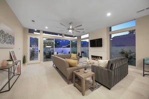 A modern living room with a fireplace, large windows, and a ceiling fan.